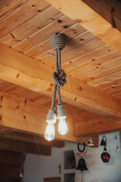 Wisząca lampa z dwiema żarówkami owinięta grubym sznurem, na tle drewnianego stropu